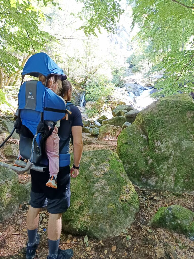 Randonnée en famille : un randonneur vu de dos porte un jeune enfant dans un porte-bébé de randonnée bleu et gris sur un sentier forestier. Ils observent une cascade au milieu de gros rochers couverts de mousse verte, dans une atmosphère paisible et lumineuse.